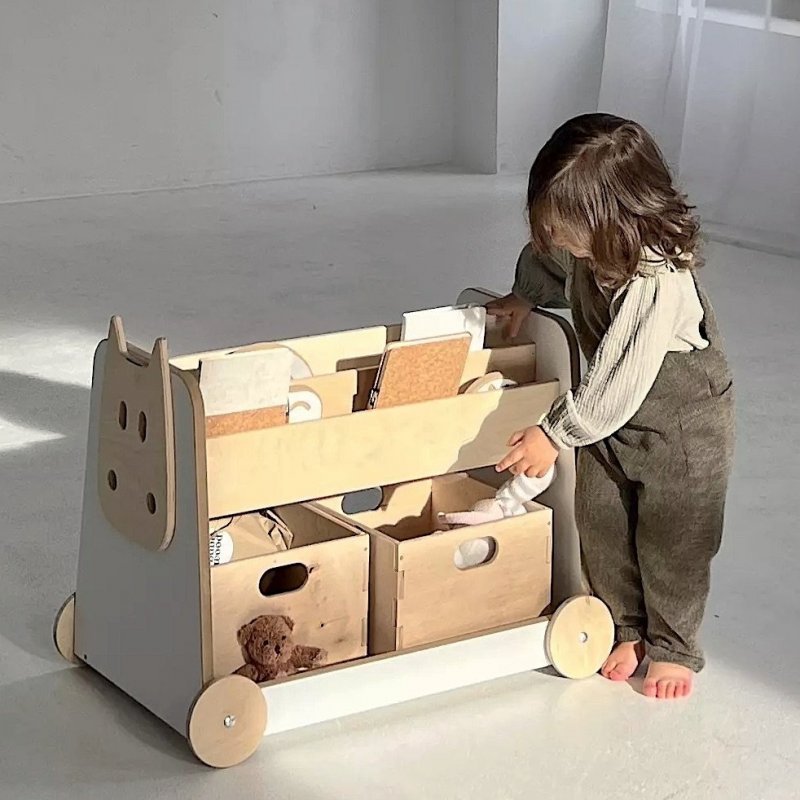 Kleines Kind mit weißem Bücherregal auf Rädern und zwei Spielzeugkisten im Kinderzimmer